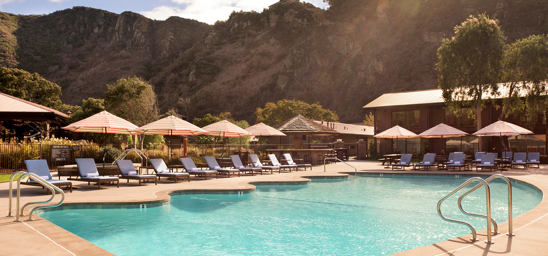The saline pool at midday, loungers and cream umbrellas set around it and the low timber pool house behind, with the canyon ridge rising steeply beyond.